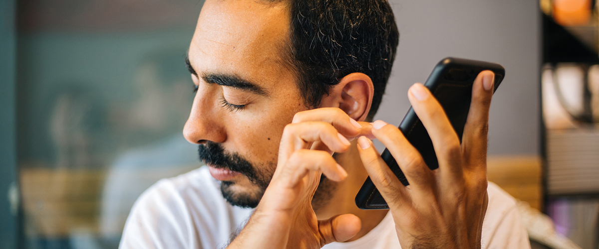 A man with brown skin, salt and pepper hair, and facial hair holds his smartphone to his ear and turns his face away listening to his phone’s screen reader.
