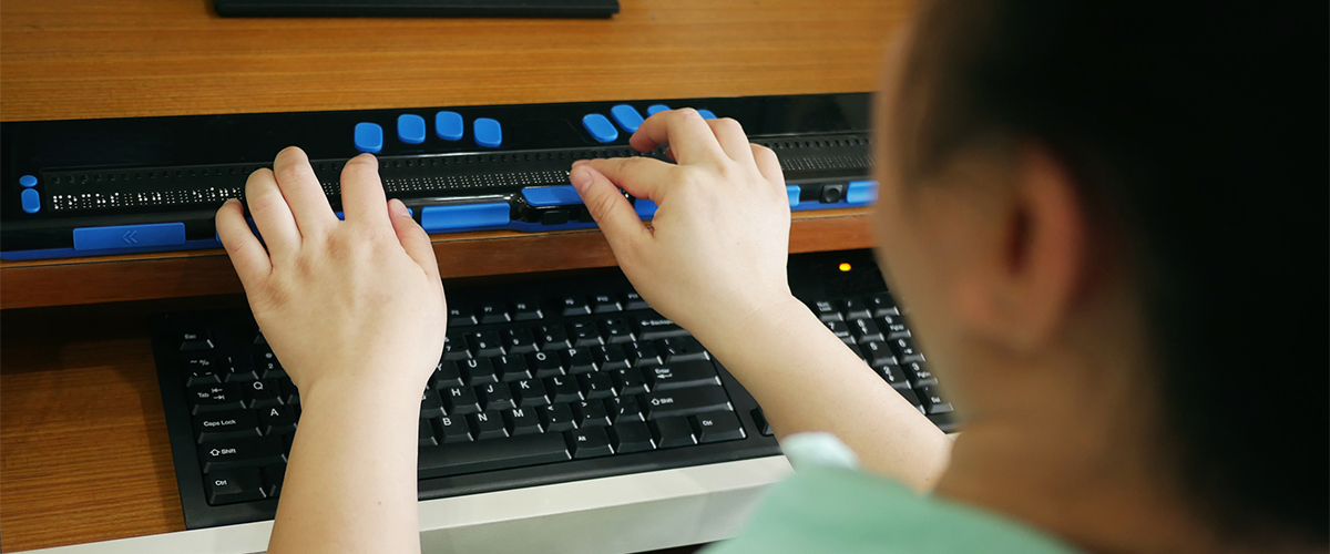 A view from behind a woman with dark hair wearing a green shirt sitting at a braille keyboard ready to type.