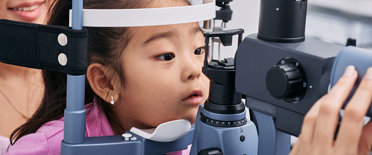 A young girl with light brown skin and dark hair in a ponytail is having her eyes examined at the eye doctor using a specialized examination tool. Her chin is placed in a chin resting area with her forehead pushed against the head guard. 